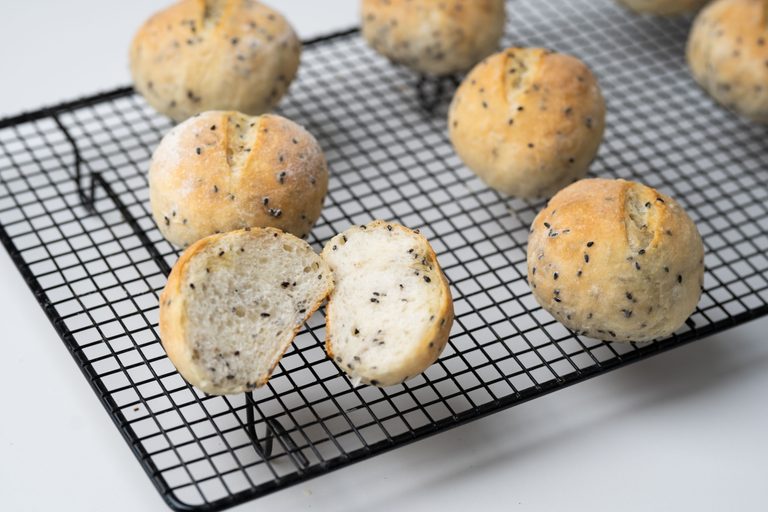 Seeded bread rolls cooling on a black wire rack, one torn open to show the crumb