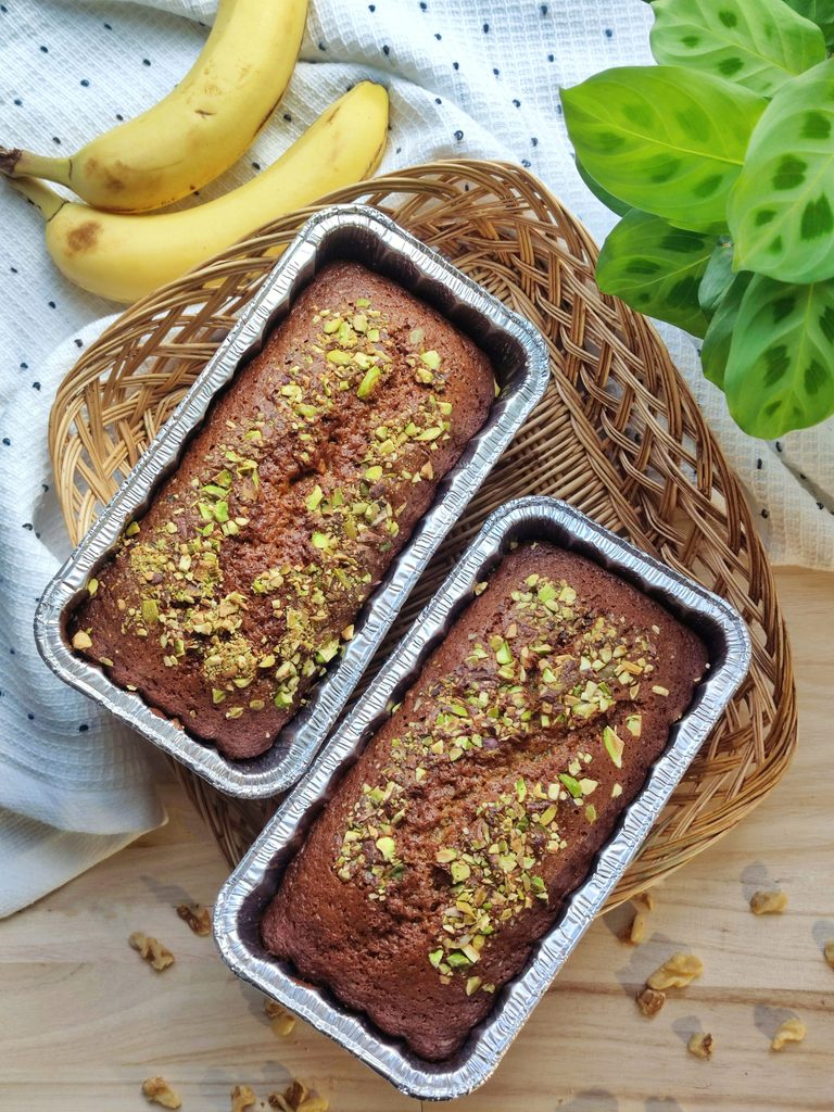Two banana bread loaves in foil pans topped with chopped pistachios, in a wicker basket